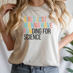 以下が含まれることがあります： 虹色の文字で「Girls Just Wanna Have Funding For Science」と書かれた白いTシャツです。