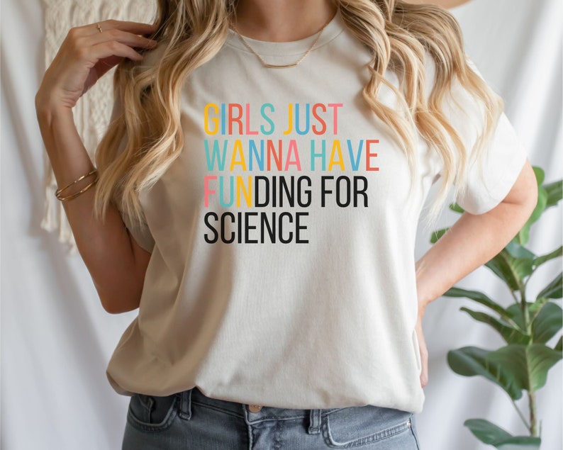 以下が含まれることがあります： 虹色の文字で「Girls Just Wanna Have Funding For Science」と書かれた白いTシャツです。