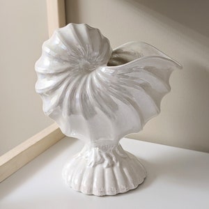 May include: A white ceramic seashell vase with a pearlescent finish. The vase is shaped like a nautilus shell and has a wide opening at the top. It sits on a pedestal base.