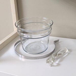 May include: A clear glass planter with a metal ring and a glass handle. The planter has a double rim and is perfect for displaying plants or flowers.