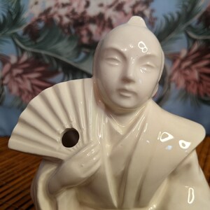 May include: White ceramic figurine of a person holding a fan. The figurine has a hole in the fan for pouring liquid.