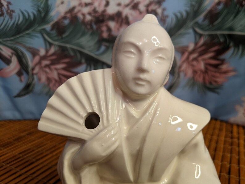 May include: White ceramic figurine of a person holding a fan. The figurine has a hole in the fan for pouring liquid.