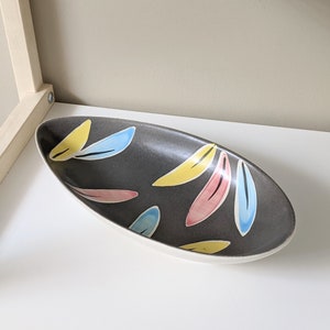 May include: A gray oval serving dish with a white rim and a colorful abstract design. The design features yellow, pink, and blue leaf shapes.