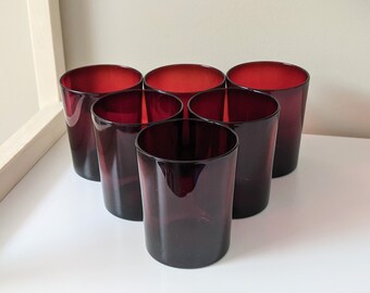 May include: Six dark red glass tumblers arranged in a pyramid shape on a white surface.