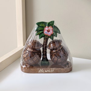 May include: Salt and pepper shakers shaped like coconuts with a wooden palm tree stand. The shakers are brown and have a woven pattern. The stand has a pink hibiscus flower with the word "Hawaii" on it.