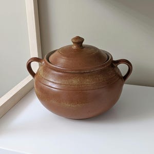 May include: A brown ceramic casserole dish with a lid and two handles. The dish has a speckled glaze and a rounded shape.