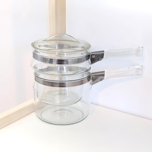 May include: Clear glass double boiler with a lid and two handles. The glass pots are held together with silver metal bands. The handles are clear plastic. The double boiler is designed for cooking and is a kitchen essential.