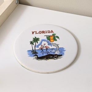 May include: White ceramic coaster featuring a colourful illustration of a Florida scene. The coaster depicts a palm tree, two pink flamingos, a dolphin leaping from the water, an alligator, and the word "FLORIDA" in red lettering.