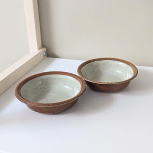 May include: Two small brown ceramic bowls with a white speckled glaze inside. The bowls are sitting on a white surface.