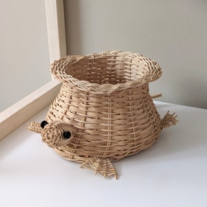 May include: A woven wicker basket shaped like a turtle with black eyes. The basket is light brown and has a wide opening at the top.