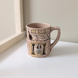May include: A brown ceramic mug with a fireplace design. The mug has a handle and the word "MUG" written on the front.