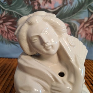 May include: White ceramic figurine of a woman with her hair pulled back in a bun. She is wearing a traditional Japanese kimono and has a serene expression on her face.
