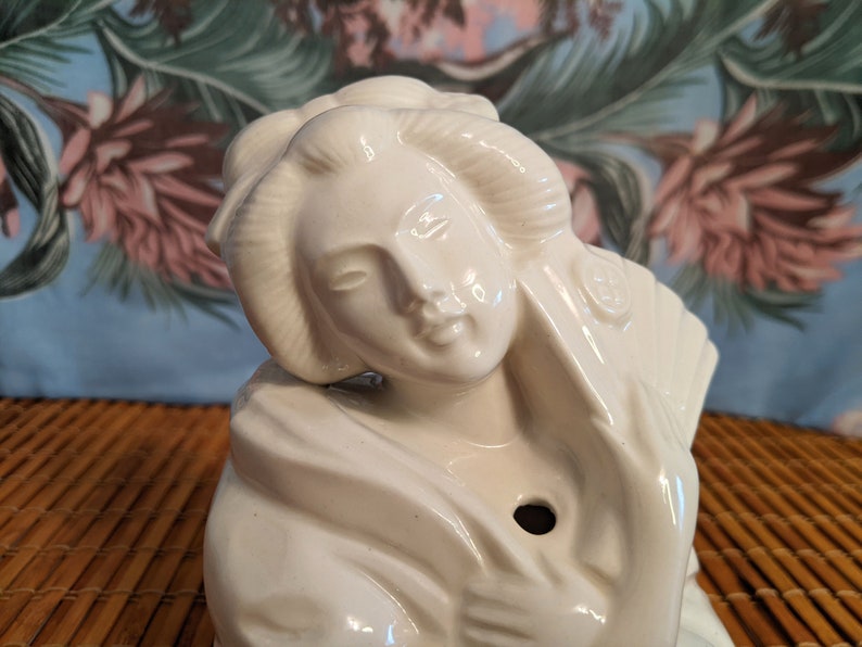 May include: White ceramic figurine of a woman with her hair pulled back in a bun. She is wearing a traditional Japanese kimono and has a serene expression on her face.