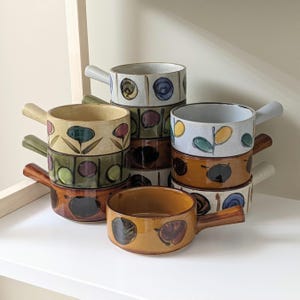 May include: A set of eight vintage ceramic soup bowls with handles. The bowls are decorated with various colourful patterns, including flowers, leaves, and geometric shapes. The bowls are stacked on top of each other, with the handles facing outwards.