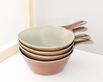 May include: A stack of four ceramic bowls with handles. The bowls have a light brown exterior and a cream-colored interior. The handles are a darker brown. The bowls are stacked on top of each other.
