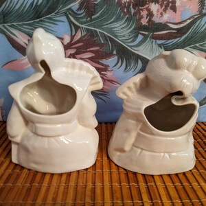 May include: Two white ceramic planter figurines in the shape of people. The figurines are facing away from the camera and have a hole in the back for planting.