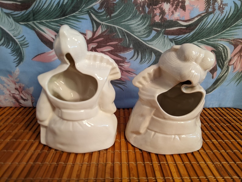 May include: Two white ceramic planter figurines in the shape of people. The figurines are facing away from the camera and have a hole in the back for planting.