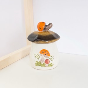 May include: A ceramic mushroom-shaped jar with a lid. The lid is brown and black, topped with an orange ladybug. The jar is white with a painted mushroom design in orange, red, and green. The jar is on a white surface.