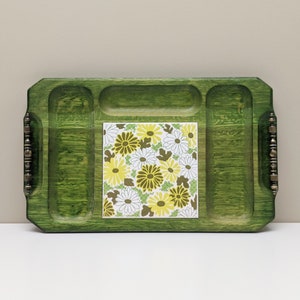 May include: A green wooden serving tray with three compartments and a removable tile insert featuring a floral pattern in shades of yellow, white, and brown. The tray has brass handles on each side.