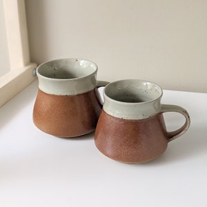 May include: Two brown and white ceramic mugs with a flared shape and a handle. The mugs have a light brown glaze on the bottom and a white glaze on the top.