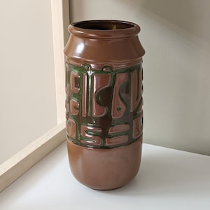 May include: A brown ceramic vase with a geometric pattern in shades of green and brown. The vase has a wide mouth and a rounded base.