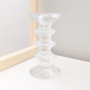Peut inclure: Un bougeoir en verre transparent présentant une surface texturée et givrée avec un design à gradins distinct. L'objet possède une large base circulaire, une colonne centrale composée de trois crêtes arrondies empilées et un plateau supérieur plat.