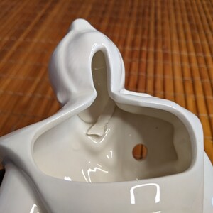 May include: White ceramic planter shaped like a person with a hooded head. The planter has a hole in the bottom for drainage and a small opening in the back for hanging.
