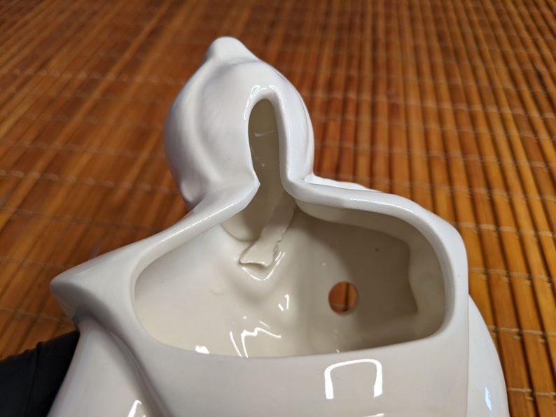 May include: White ceramic planter shaped like a person with a hooded head. The planter has a hole in the bottom for drainage and a small opening in the back for hanging.