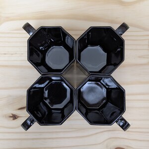 May include: Four black octagonal mugs with handles, arranged in a square pattern on a wooden surface.