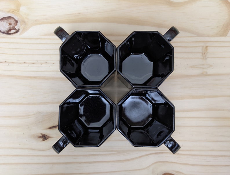 May include: Four black octagonal mugs with handles, arranged in a square pattern on a wooden surface.