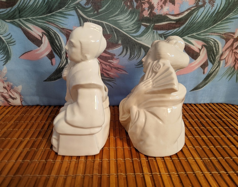 May include: Two white ceramic figurines of people in traditional Japanese clothing. The figurines are sitting on a woven mat.