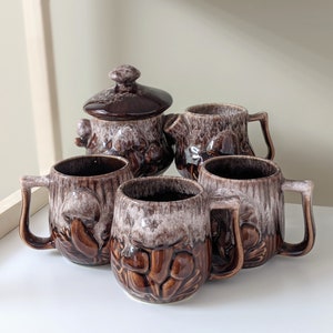 May include: A set of brown and white ceramic mugs with mushroom designs. The set includes a teapot with a matching design.