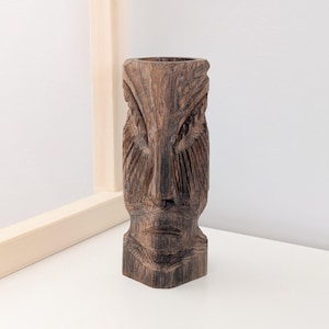 May include: A hand-carved wooden tiki statue with a detailed face and textured surface. The brown sculpture has a cylindrical shape with a hollowed-out top, likely for holding items. The artwork is displayed on a white surface.