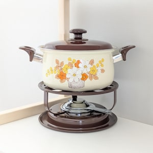 May include: Cream enamel pot with a brown lid and handles, decorated with a floral pattern in yellow, orange, and white. The pot rests on a brown metal stand with a burner and spoon. A vintage kitchen item.