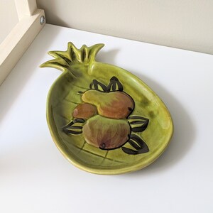 May include: A green ceramic pineapple-shaped dish with a painted design of pears. The dish is perfect for serving fruit or snacks.