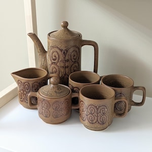 May include: A brown and cream ceramic teapot and four matching cups with a swirling pattern. The teapot has a handle and a spout. The cups have handles. The set is on a white surface.