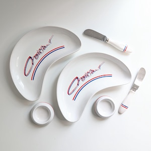 May include: A white ceramic croissant plate set with two plates, two small dishes, and two butter knives. Each plate has the word "Croissant" in red and blue, with matching stripes. The knives and dishes have red and blue accents.