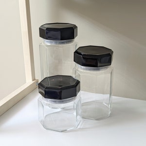 May include: Three glass canisters with black octagonal lids. The canisters are clear glass and are arranged in a row from largest to smallest.