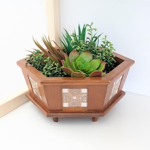 May include: A wooden hexagonal planter filled with a variety of faux succulents and greenery. The planter is a warm brown color with decorative tile inserts on the sides. The arrangement includes a large, green succulent in the center.