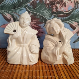 May include: Two white ceramic figurines of people in traditional Japanese clothing. The figurine on the left is holding a fan and the figurine on the right is holding a flower. Both figurines have a hole in the top of their heads, suggesting they are used as drink holders.