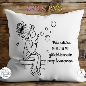 May include: White decorative pillow with a black and white illustration of a girl blowing bubbles. The pillow has the text "Wir sollten mehr Zeit mit glücklichsein verplempern". The pillow is on a wooden surface.