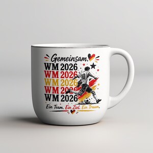May include: White ceramic mug with a soccer theme. The mug features the text "Gemeinsam" and "WM 2026" in red, yellow, and black, along with a soccer player graphic. The phrase "Ein Team. Ein Ziel. Ein Traum." is also included.