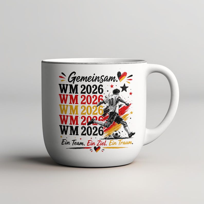 May include: White ceramic mug with a soccer theme. The mug features the text "Gemeinsam" and "WM 2026" in red, yellow, and black, along with a soccer player graphic. The phrase "Ein Team. Ein Ziel. Ein Traum." is also included.