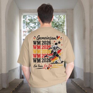 May include: A tan t-shirt with a graphic design featuring the text "Gemeinsam" and "WM 2026" in red, yellow, and black. A soccer player is kicking a ball. The text "Ein Team. Ein Ziel. Ein Traum." is also present.