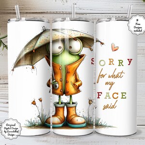 Puede incluir: Vaso blanco con una ilustración caprichosa de una rana. La rana lleva un impermeable y botas naranjas, sosteniendo un paraguas. El texto en el vaso dice "Sorry for what my FACE said." Incluye una pajita y un corazón.