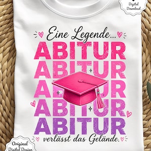 Könnte beinhalten: Weißes T-Shirt mit einem Abschluss-Thema. Das Design zeigt das Wort "ABITUR" in Pink und Lila, eine Abschlusskappe und deutschen Text. Das Shirt wird auf einer gewebten Oberfläche präsentiert.