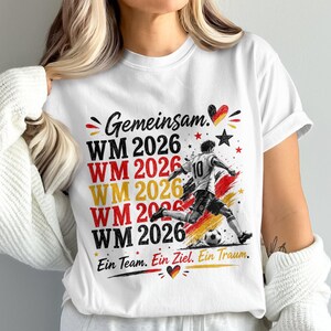 May include: White t-shirt with a soccer-themed graphic. The design includes the text "Gemeinsam WM 2026" in red, yellow, and black, along with a soccer player kicking a ball. The shirt also has the text "Ein Team. Ein Ziel. Ein Traum."