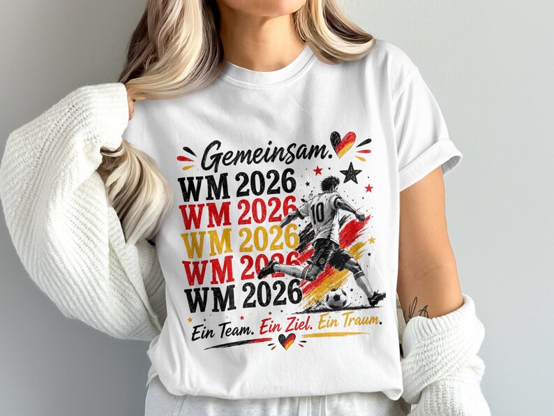 May include: White t-shirt with a soccer-themed graphic. The design includes the text "Gemeinsam WM 2026" in red, yellow, and black, along with a soccer player kicking a ball. The shirt also has the text "Ein Team. Ein Ziel. Ein Traum."