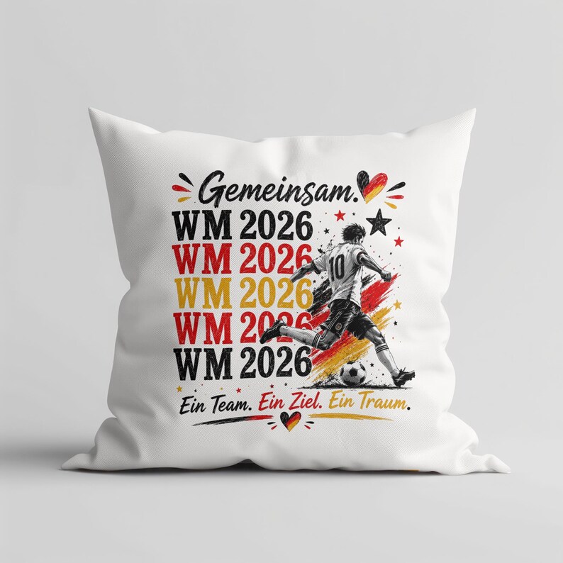 May include: White square pillow with a soccer theme. The pillow features the text "Gemeinsam. WM 2026" in red, yellow, and black, along with a graphic of a soccer player kicking a ball. The phrase "Ein Team. Ein Ziel. Ein Traum." is also included.