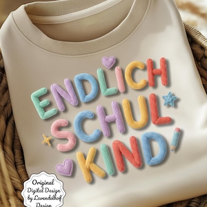 May include: Beige sweatshirt with the text "ENDLICH SCHUL KIND" in colorful, textured letters. The design includes a star, a pencil, and heart shapes. The sweatshirt has a round neckline and is made of a soft material.